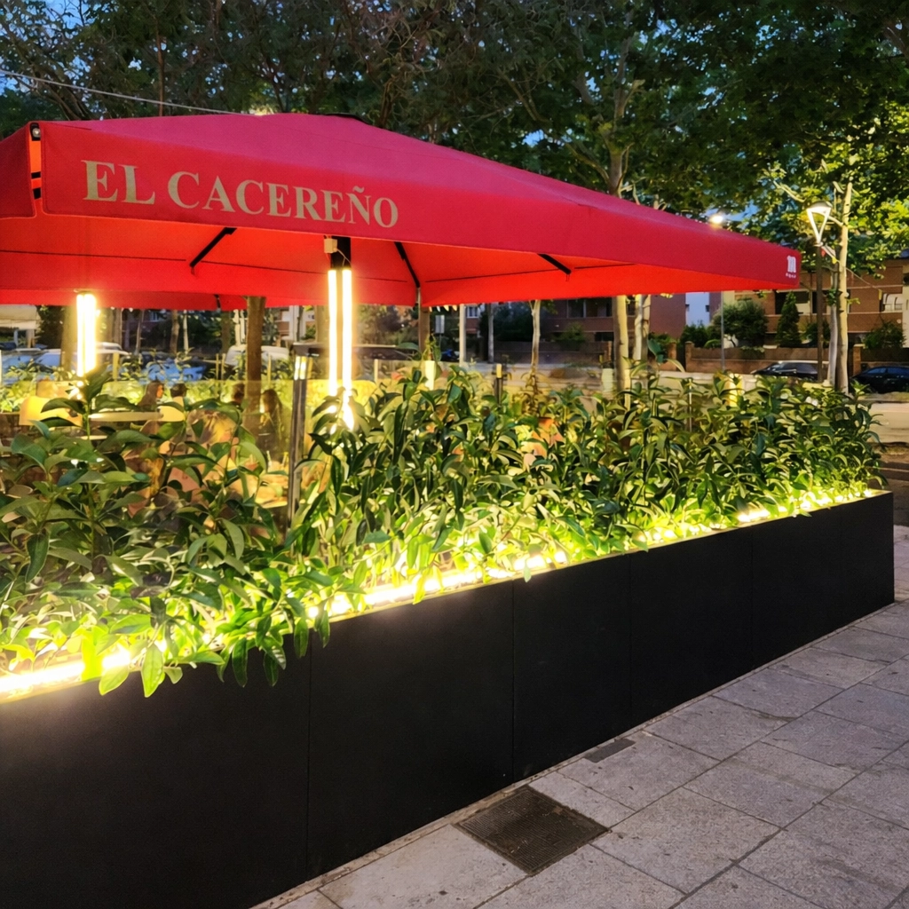 Terraza de restaurante con parasol y jardineras iluminadas al anochecer